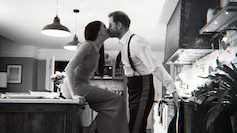 Black and white photograph of Meghan sat on a kitchen counter in a gown and Harry in a suit without his jacket leaning in to kiss her.