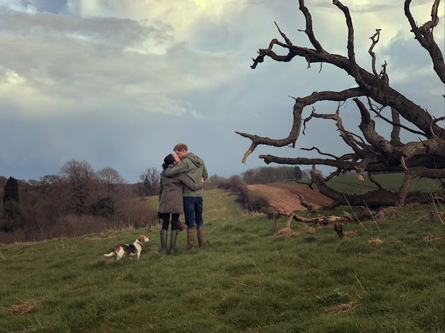 Harry and Meghan photographed from behind, embracing on a dog walk in expansive fields, wearing matching green coats.